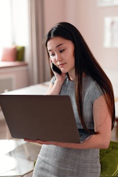 Asian woman multitasking on phone and laptop indoors. Modern work scene.