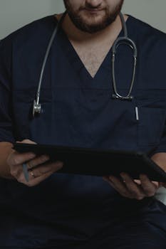 Doctor in navy scrubs with stethoscope, holding a digital tablet, focuses on patient information.