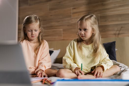 Two young girls sitting indoors learning online with a laptop in a cozy home setting, focused and engaged.